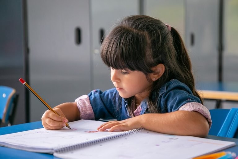 طفلة يجلس على مكتب ويكتب في دفتر بمساعدة قلم رصاص. يظهر الطفل مركزًا على المهمة بجدية - دليل شامل لتعليم الحروف العربية للأطفال في المنزل